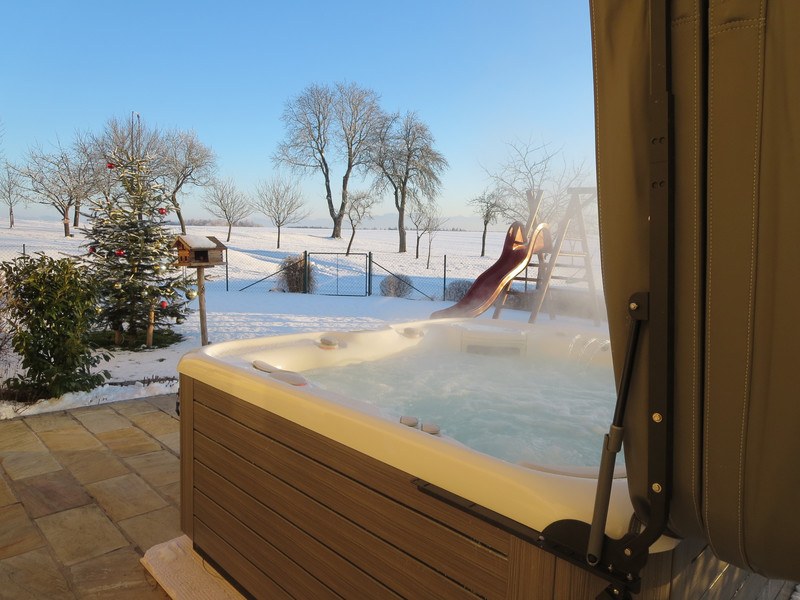A steaming hot tub with opened cover on a snow-covered patio with a view of a winter landscape