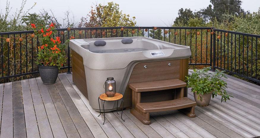 Brown outdoor hot tub on a wooden deck with small steps, railing, and potted plants nearby at dusk or overcast day.