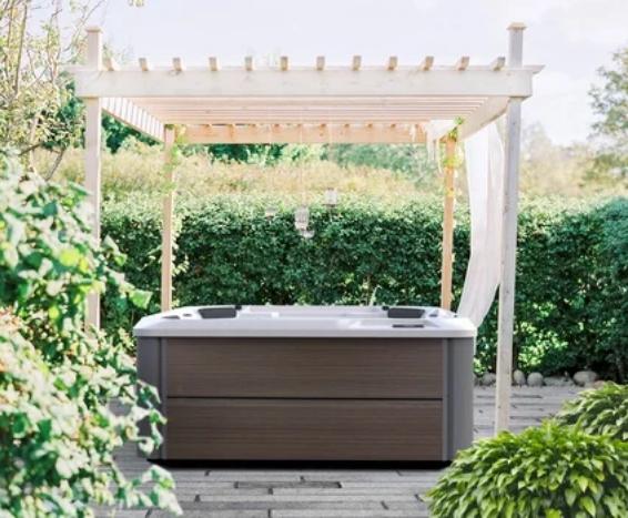 Outdoor hot tub beneath a white pergola in a sunny garden, with green hedges around it.